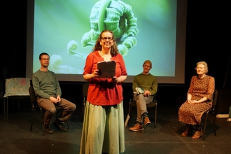 3 performers sat on a stage with one stood in the front centre of the stage reading a poem 