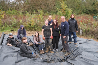 8 people stood in a dug out pond area with a pond liner in