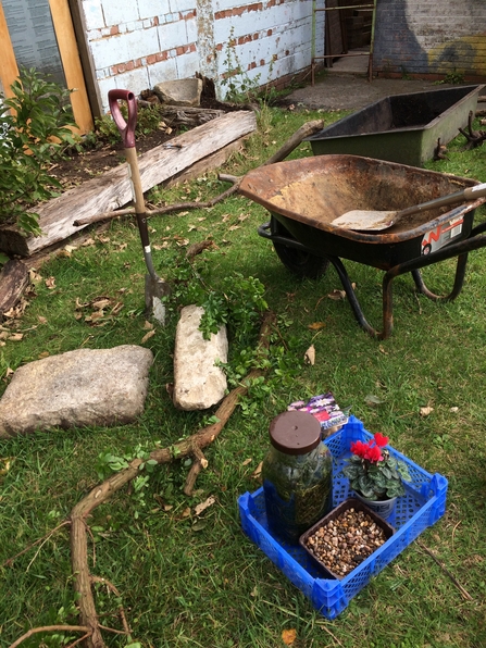 Pond building tools on ground. Spade in ground. Wheelbarrow in the corner with spade in it. Box of pond plants in the other corner.