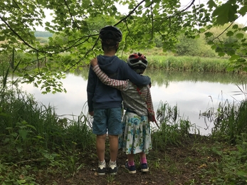 Henry and Elsie, wearing cycle helmets, embrace while looking over the water