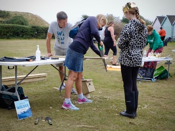 Woman sawing on wood at the end of table as someone else helps out and child looks on.