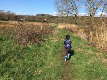 Boy running on footpath in between fields. Bright blue skies, but not leaves on trees.