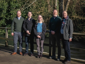 5 people line up for a photo along a fence by a bank of a chalk stream 