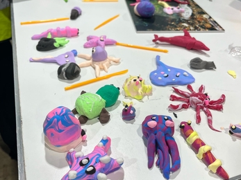 A table covered with various colorful clay models created by attendees at a crafting workshop, including marine animals and abstract shapes, with visible crafting tools and instruction materials.