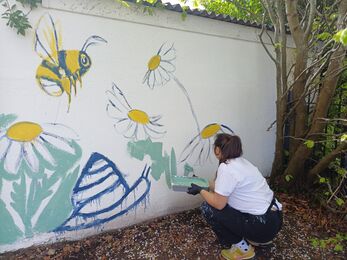 a lady in the process of painting a large wall mural with daisies and bumblebees