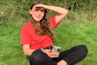Finola in a red shirt and black trousers, sitting on the grass, smiling at the camera as she shades her eyes from the sun with her hand.