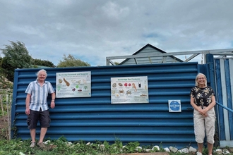 Two people stood against a blue fence with two interpretation panels showcasing lots of marine wildlife on them. 