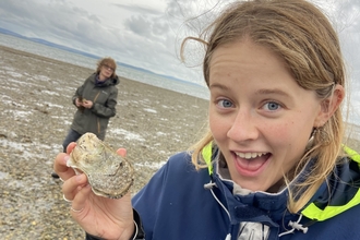 A person (Milly) wearing a jacket excitedly showing a shell on a beach, with another person visible in the background.