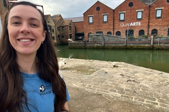 A person smiling in the foreground with the Quay Arts centre and a calm water canal in the background.