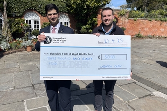 2 men presenting a large cheque to Hampshire & Isle of Wight Wildlife Trust for £3000. They're standing in front of a bricked building