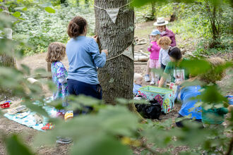 Children playing in a woodland with bunting round a tree trunk and a picnic blanket on the ground.