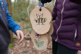 Two wooden log slices with a bee and flower picture on, held by children playing in a woodland.