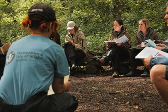 A woman wearing a Hamsphire & Isle of Wight Wildlife Trust branded blue top facing a group of adults in an outdoor classroom environment