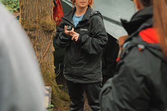 A member of Trust staff standing in front of outdoor shelters explaining to a group of adults