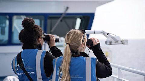 Two women wearing blue high vis jackets, looking out of binoculars overlooking the sea