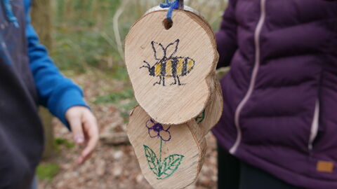 Two wooden log slices with a bee and flower picture on, held by children playing in a woodland.