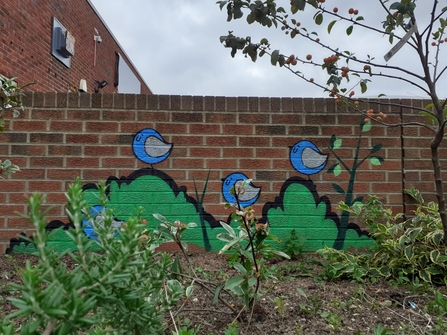 View of raised bed with plants. In the back there's a brick wall with a mural of birds, painted by FARK