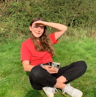 Finola in a red shirt and black trousers, sitting on the grass, smiling at the camera as she shades her eyes from the sun with her hand.