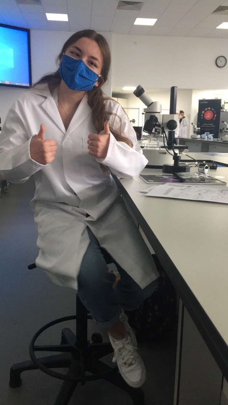 Finola in a lab, wearing a white lab coat and a face mask. Her body is turned towards the camera, away from her desk and microscope. Finola gives a big thumbs up with both hands.