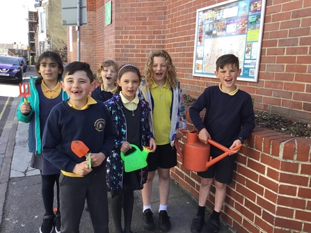 Six young pupils holding watering cans and gardening tools getting ready to do some planting