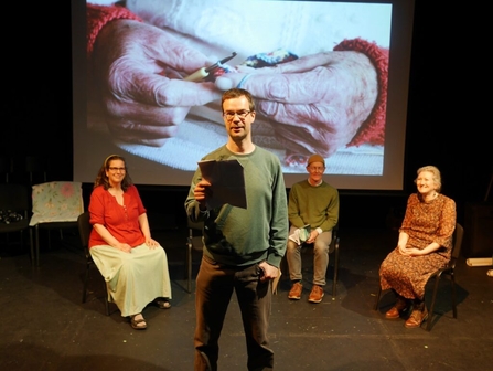 3 performers sat on a stage with one stood in the front centre of the stage reading a poem 