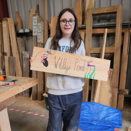 Young person in a workshop barn holding a handmade wooden sign saying wildlife pond