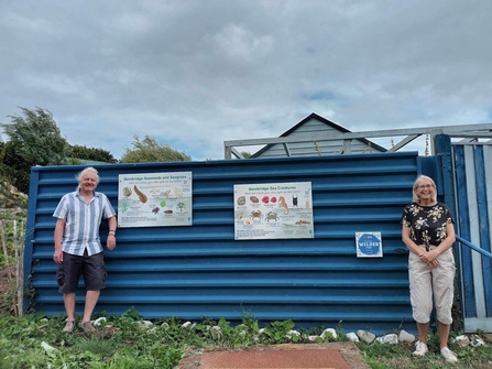 Two people stood against a blue fence with two interpretation panels showcasing lots of marine wildlife on them. 