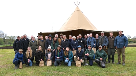 Large group of people stood and knelt in front of brown yurt tent. The people are stood on green grass. 