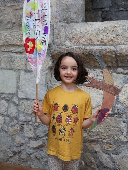 Young girl in yellow top holing a cardboard bird in her left hand and a paper banner in her right. The sign reads: "Plant more trees and flowers"