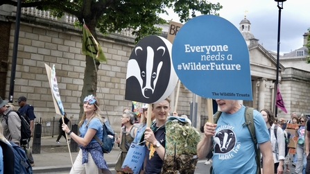 Blue cardboard sign reading 'Everyone needs a #WilderFuture next to a carboard sign of the Wildlife Trust's logo. Both are held aloft by two male Wildlife Trust staff.