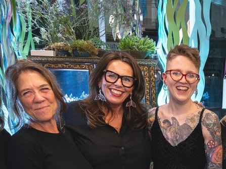 three women smiling at the camera. In the background is a variety of indoor plants.
