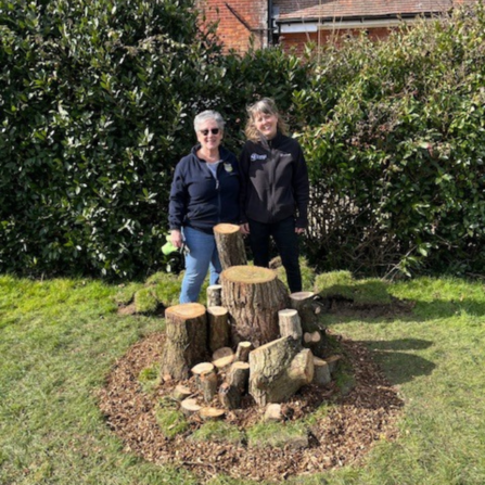 Two people stood behind a stumpery, with lush green grass surrounding them and a green hedge behind them