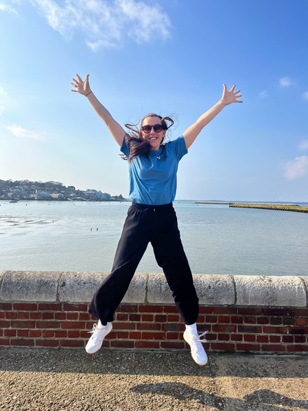 Person in a blue shirt and black trousers joyfully jumping with arms raised against a scenic backdrop with water and a clear sky.