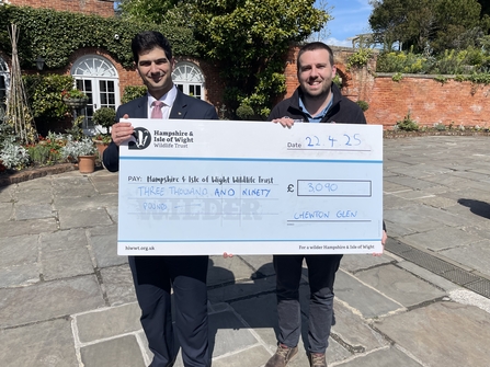 2 men presenting a large cheque to Hampshire & Isle of Wight Wildlife Trust for £3000. They're standing in front of a bricked building 