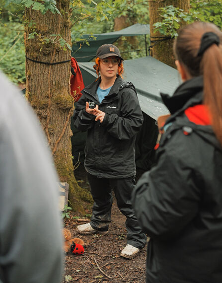 A member of Trust staff standing in front of outdoor shelters explaining to a group of adults