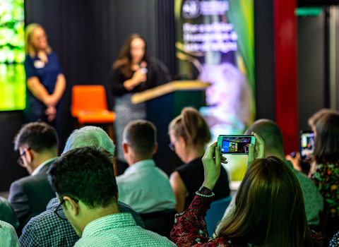 View of the back of a seated crowd facing a talker on a stage. One lady is recording the event on a raised phone