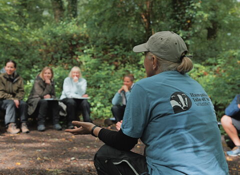 A woman wearing a Hamsphire & Isle of Wight Wildlife Trust branded blue top facing a group of adults in an outdoor classroom environment